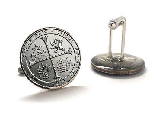 A pair of silver-colored cufflinks with an engraved design representing the San Antonio Missions National Historical Park, featuring a cross and a building.