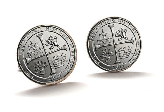 A pair of silver-colored cufflinks with an engraved design representing the San Antonio Missions National Historical Park, featuring a cross and a building.