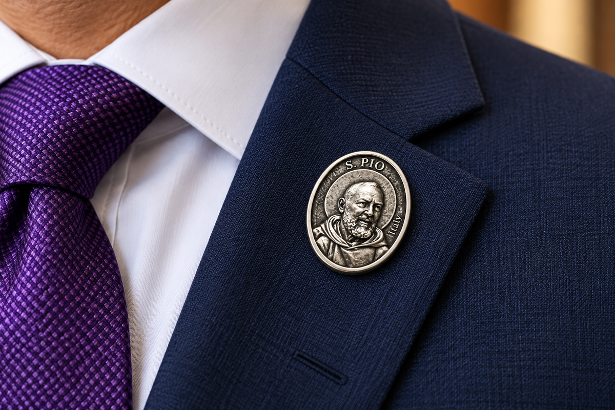 A silver-toned oval lapel pin featuring a portrait of Saint Pio of Pietrelcina, pinned to the lapel of a navy blue suit jacket worn over a white collared shirt with a purple patterned tie.