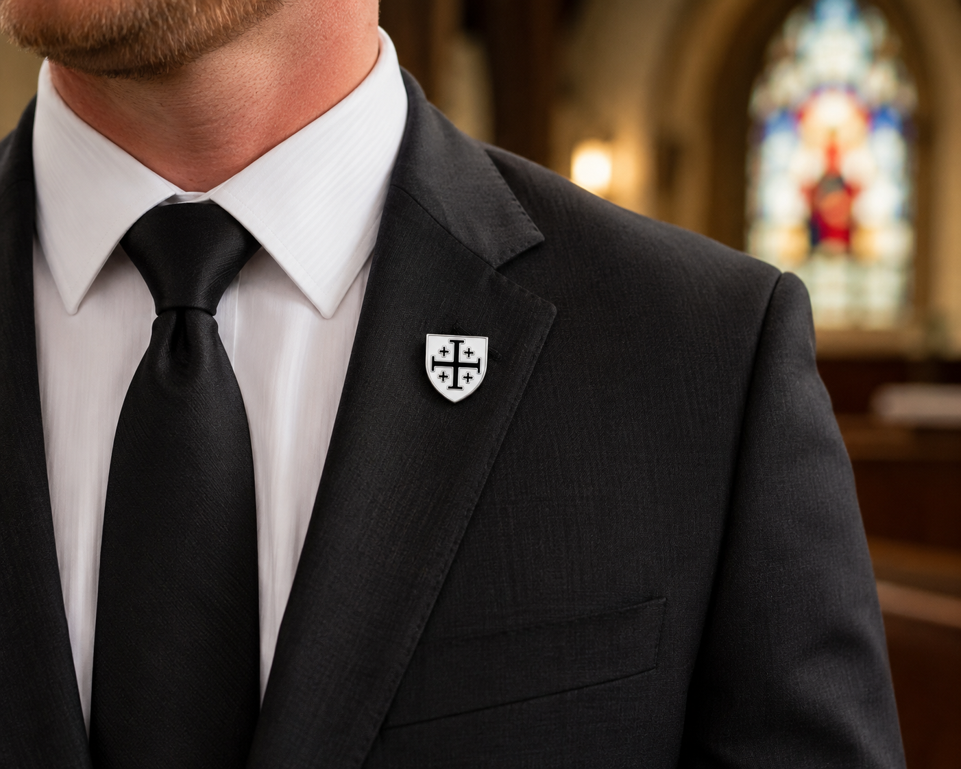 A close-up of a man wearing a black suit jacket, white dress shirt, and black tie, with a small white shield-shaped lapel pin featuring a black cross on the left lapel. The background is a softly blurred church interior with stained glass.