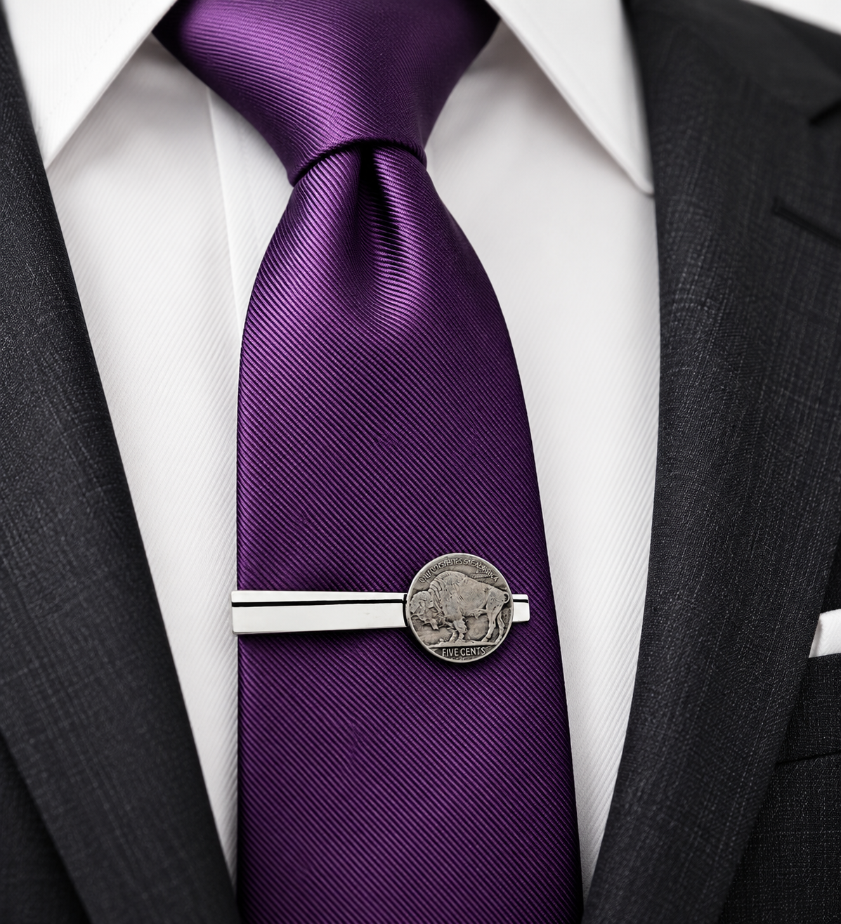 A close-up of a purple silk necktie with a silver-toned coin tie clip featuring a buffalo coin, styled on a white dress shirt and dark gray suit jacket.