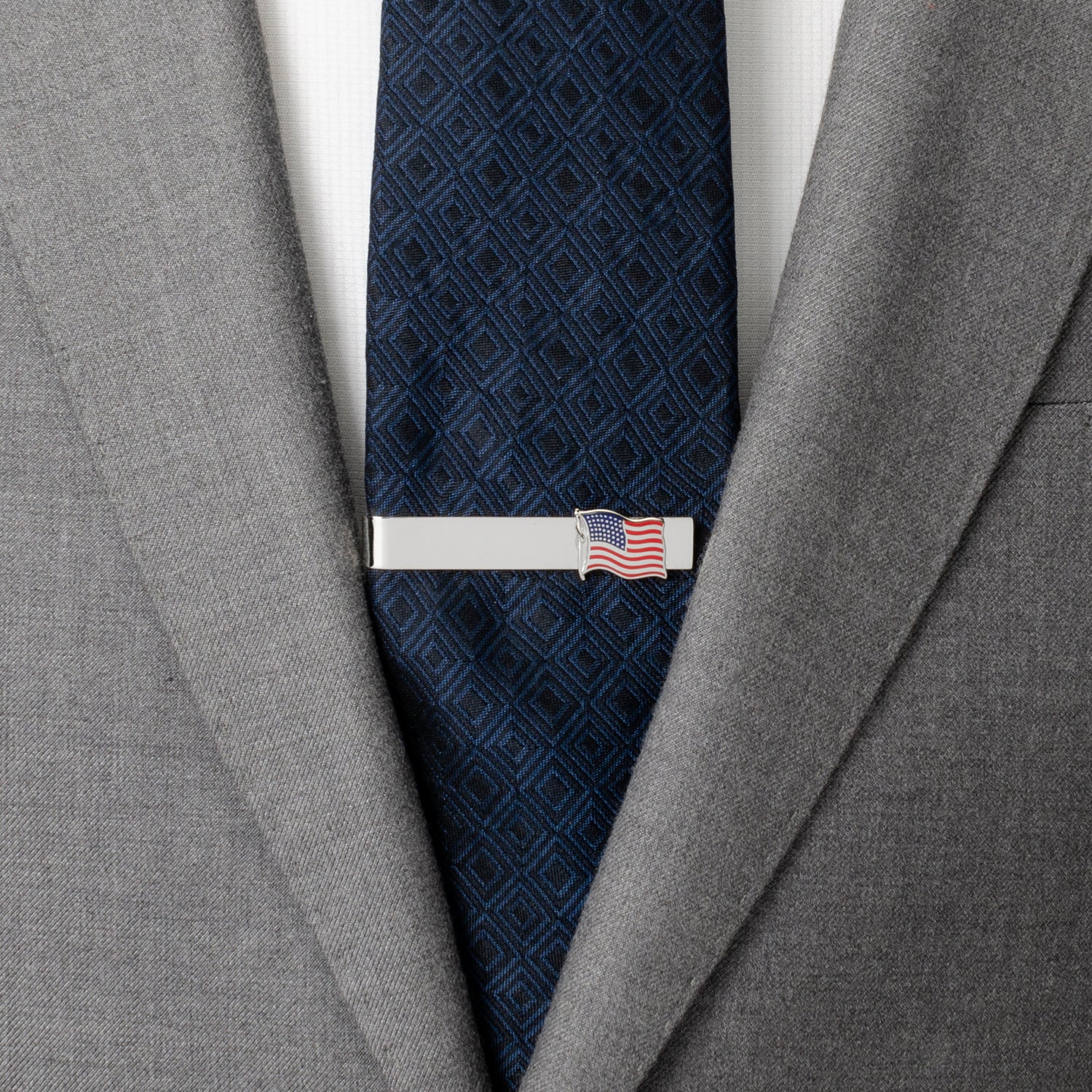 Gray suit jacket with a navy patterned tie and a silver tie clip with an American flag design.