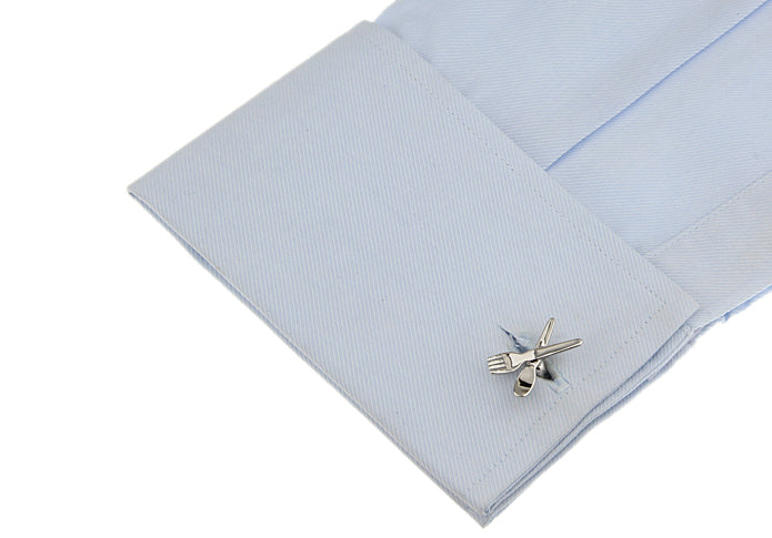 A close-up of a light blue dress shirt cuff with a silver-toned metal tie clip featuring crossed cutlery, set against a plain white background.