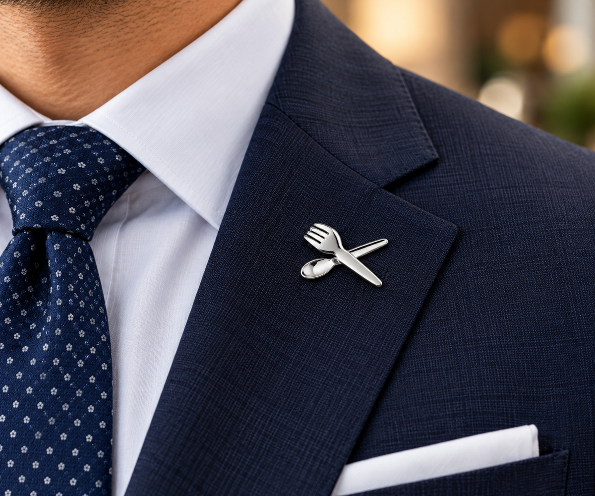 A silver-toned enamel lapel pin shaped like a crossed fork and spoon, pinned to the lapel of a man's navy blue suit jacket. The suit is styled with a white dress shirt and a navy blue patterned tie.
