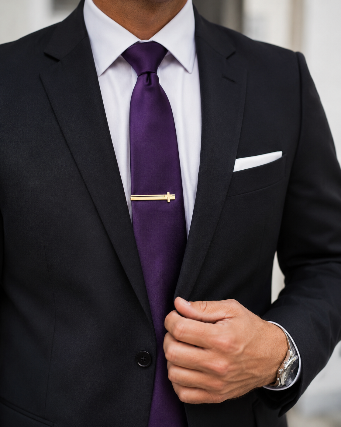 Man wearing a black suit with a purple tie and white pocket square. Gold Cross Tie Bar