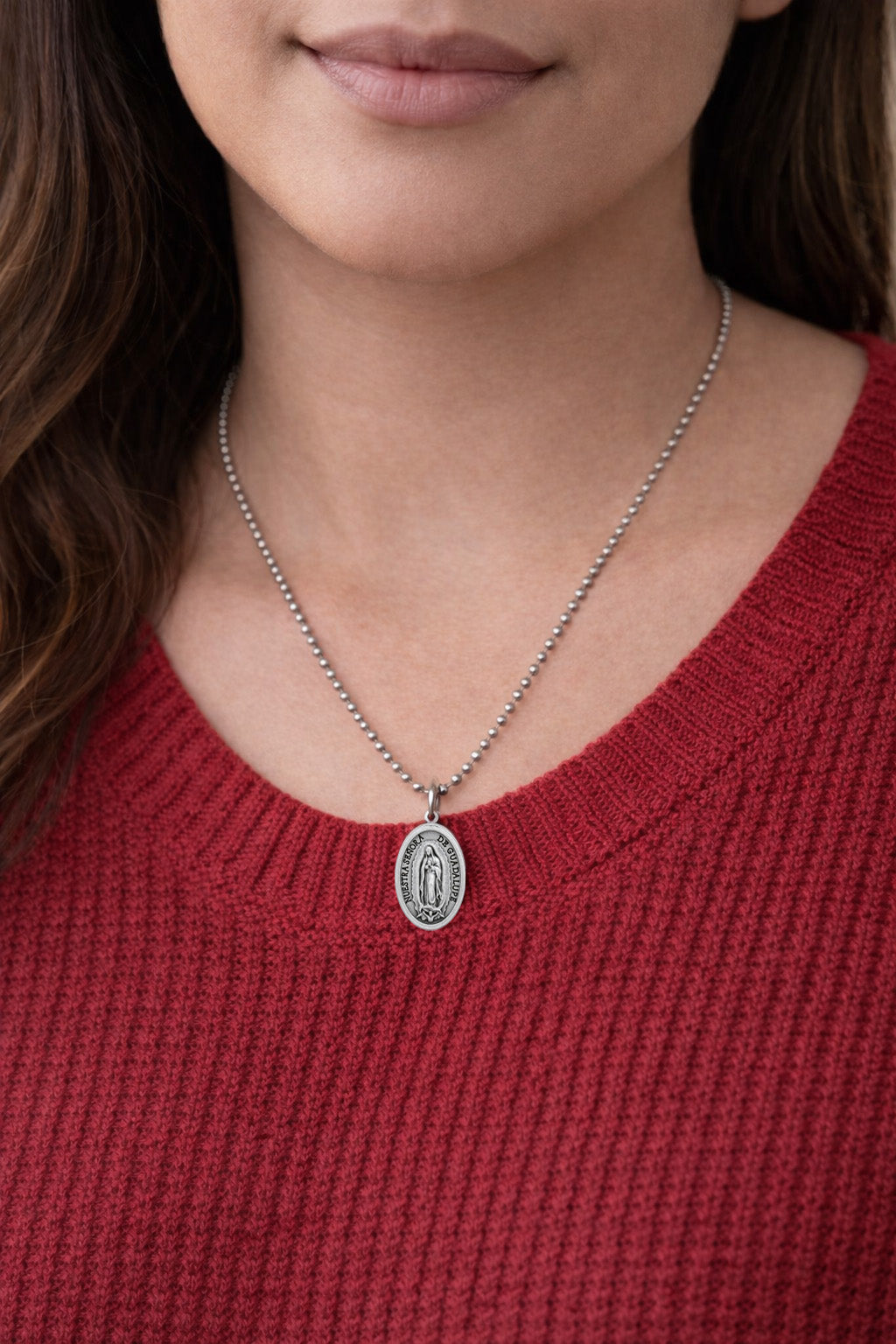 Woman wearing a silver Our Lady of Guadalupe Medal necklace with a pendant against a red sweater background