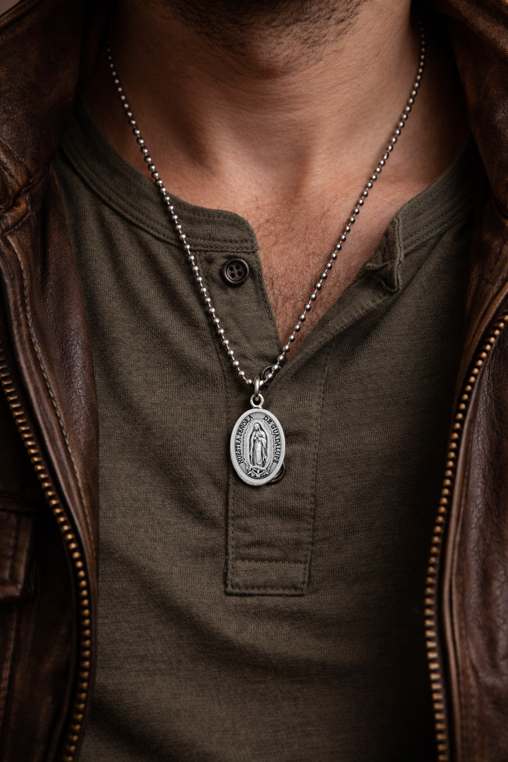 Man wearing a green shirt and brown leather jacket with a Guadalupe necklace featuring a pendant.
