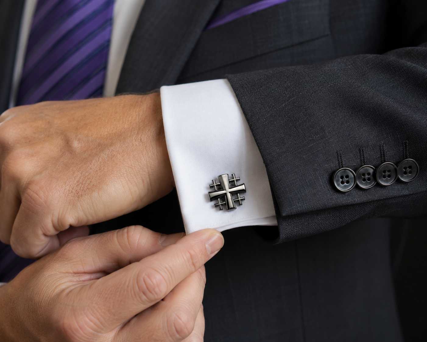 A close-up of a man's hand adjusting a silver-toned cross-shaped cufflink on a white dress shirt cuff, wearing a dark gray suit jacket and a purple striped tie.