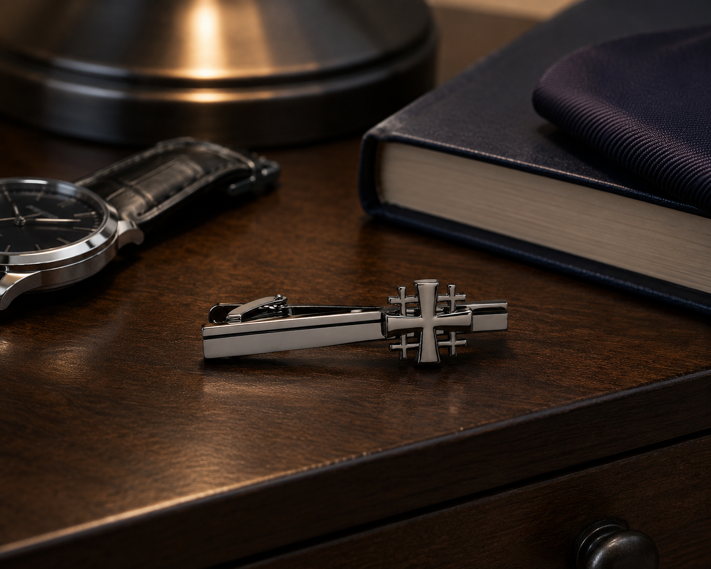 Gunmetal Jerusalem Cross Tie Clip with a cross motif resting on a dark wooden desk, surrounded by lifestyle props including a watch, a navy book, and a dark fabric accessory.