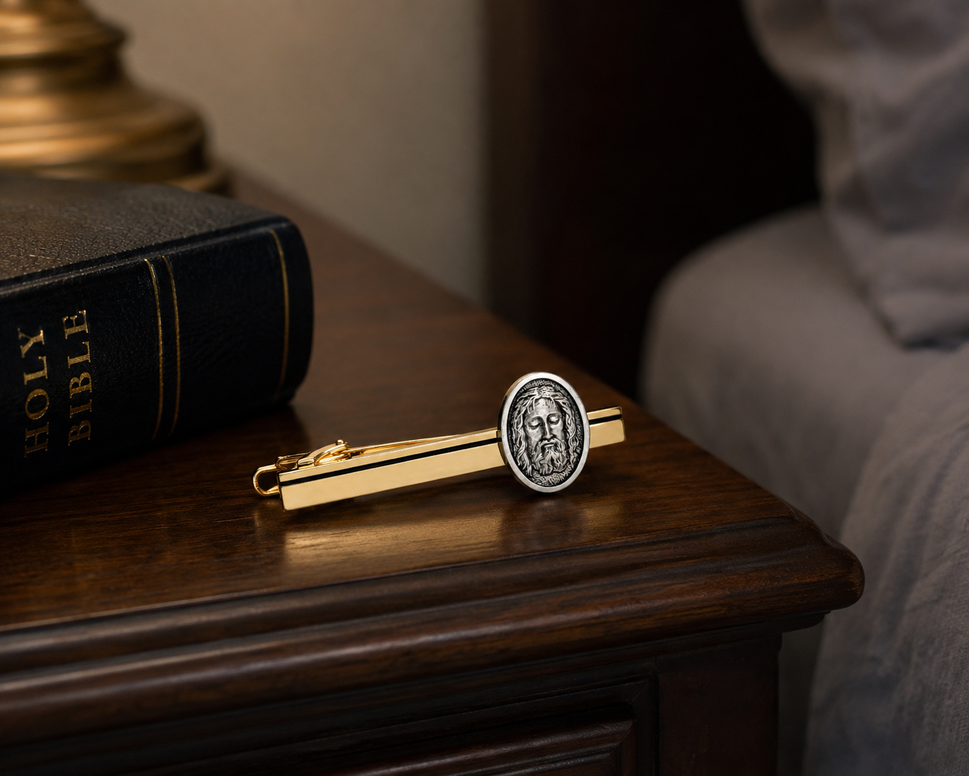 A gold-toned tie clip featuring a circular silver-toned medallion with a religious portrait, resting on a dark wooden table next to a Holy Bible.