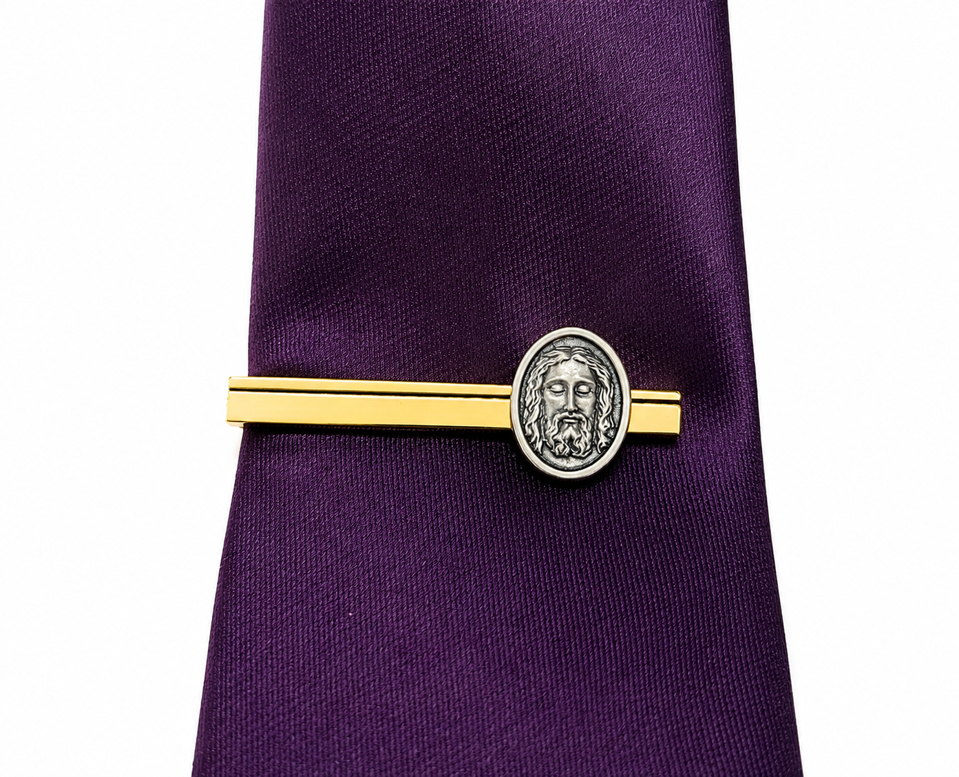 A gold tie bar with a silver oval medallion featuring a classical face, shown clipped to a purple necktie on a white background.