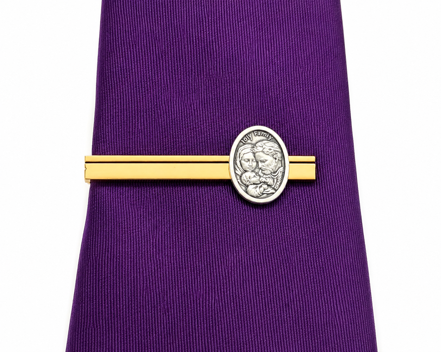 A gold tie bar with an oval silver-toned medallion featuring a lion design and the text 'Holy Family, shown tied around a purple necktie on a white background.