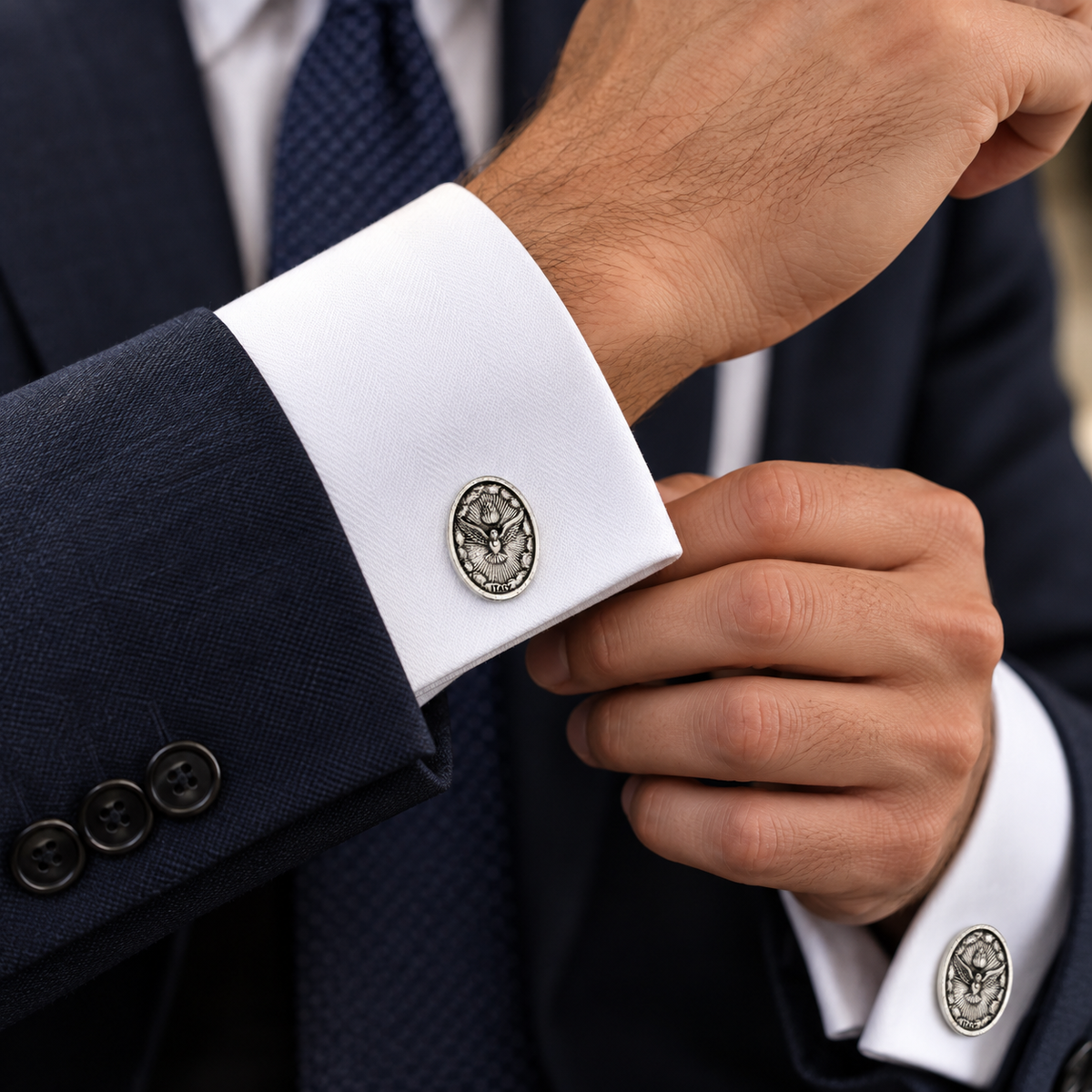 A close-up of a man's hands adjusting a white dress shirt cuff with a round silver-toned metal cufflink featuring an eagle emblem, paired with a navy suit jacket and blue tie.