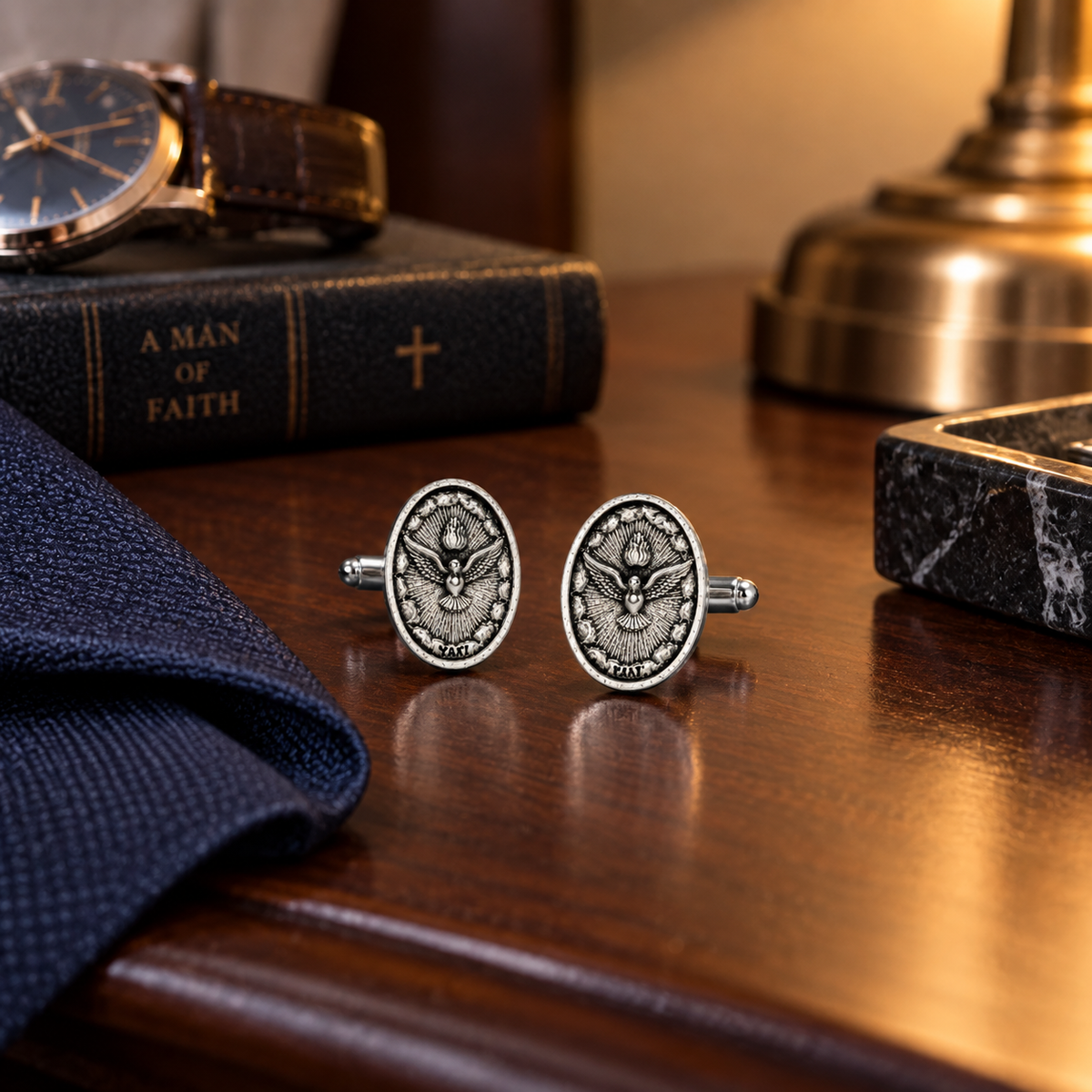 A pair of oval silver cufflinks with a detailed religious emblem, displayed on a polished wooden desk next to a navy blue tie, a vintage watch, a book, and a brass lamp base.
