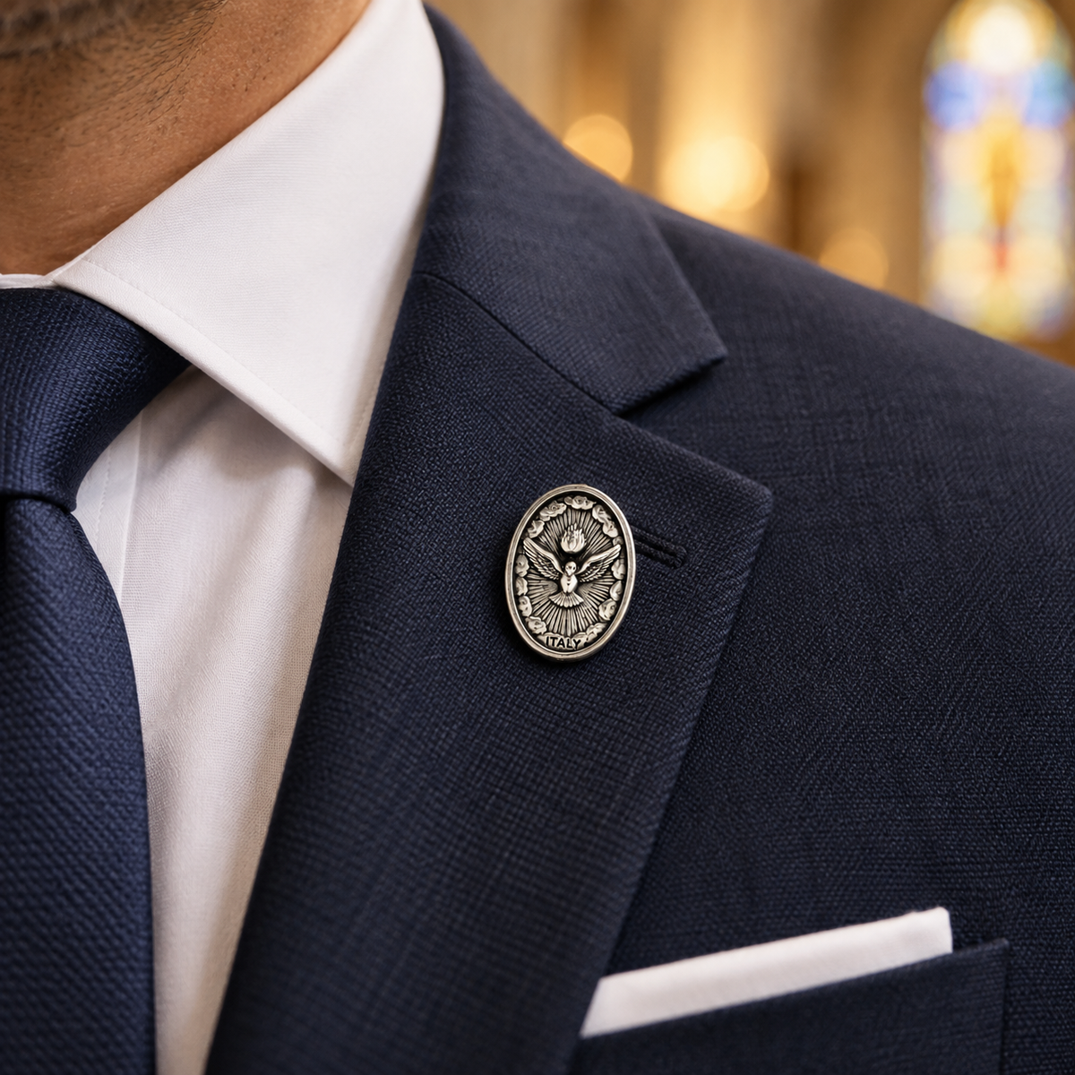 A close-up of a man wearing a navy blue suit jacket, white dress shirt, navy tie, and a white pocket square, with a silver-toned oval lapel pin featuring an eagle emblem pinned to the chest.