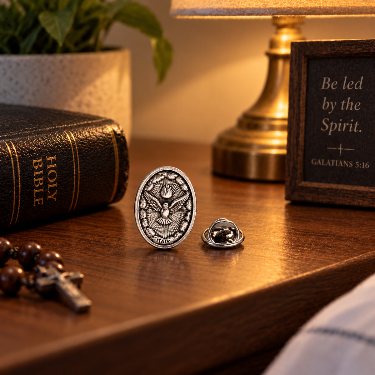 A silver-toned oval lapel pin with an eagle emblem and the words 'JESUS IS THE ONLY TRUE GOD' is displayed on a wooden table next to a Holy Bible, a framed scripture quote, a lamp, and a potted plant.