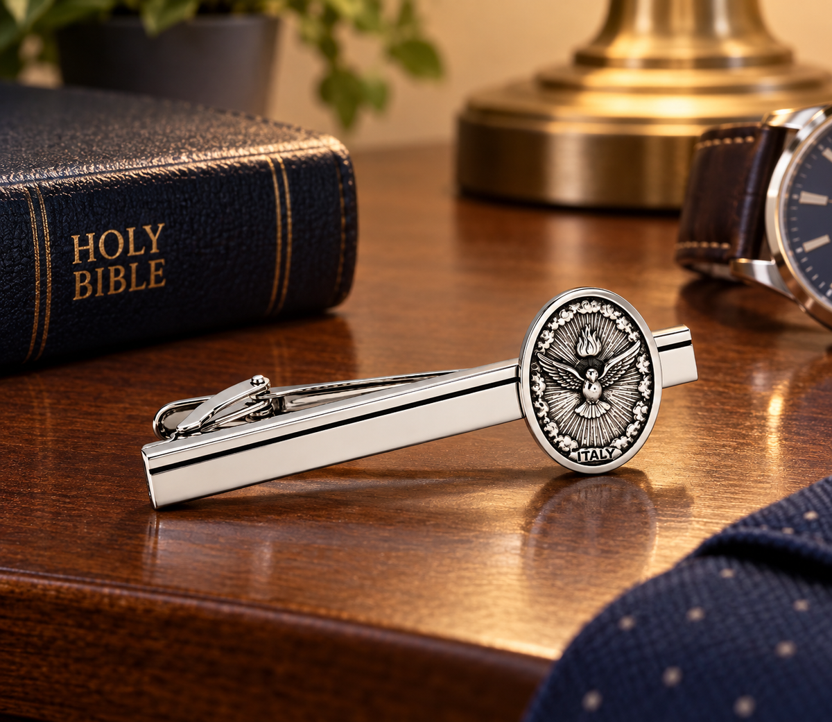 A silver-toned tie clip featuring an oval Saint Patrick emblem in the center, displayed on a polished wooden desk next to a dark leather Holy Bible, a watch, and a polka-dot necktie.