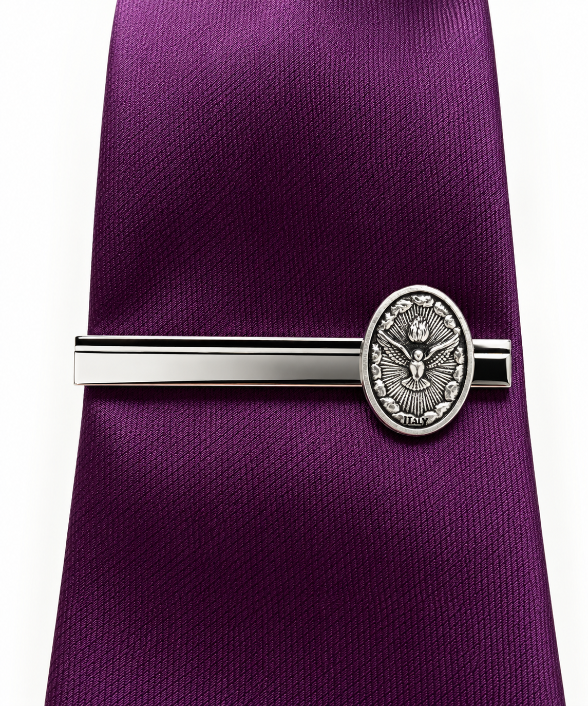 A silver-toned metal tie clip with an oval crest emblem, shown securing a textured purple necktie against a plain white background.