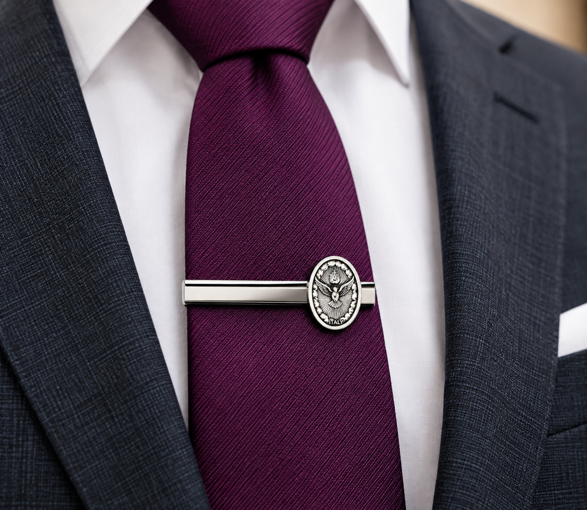 A close-up of a dark plum purple necktie with a silver-toned metal tie bar featuring an oval US Customs seal emblem, worn with a white dress shirt and a dark gray suit jacket.