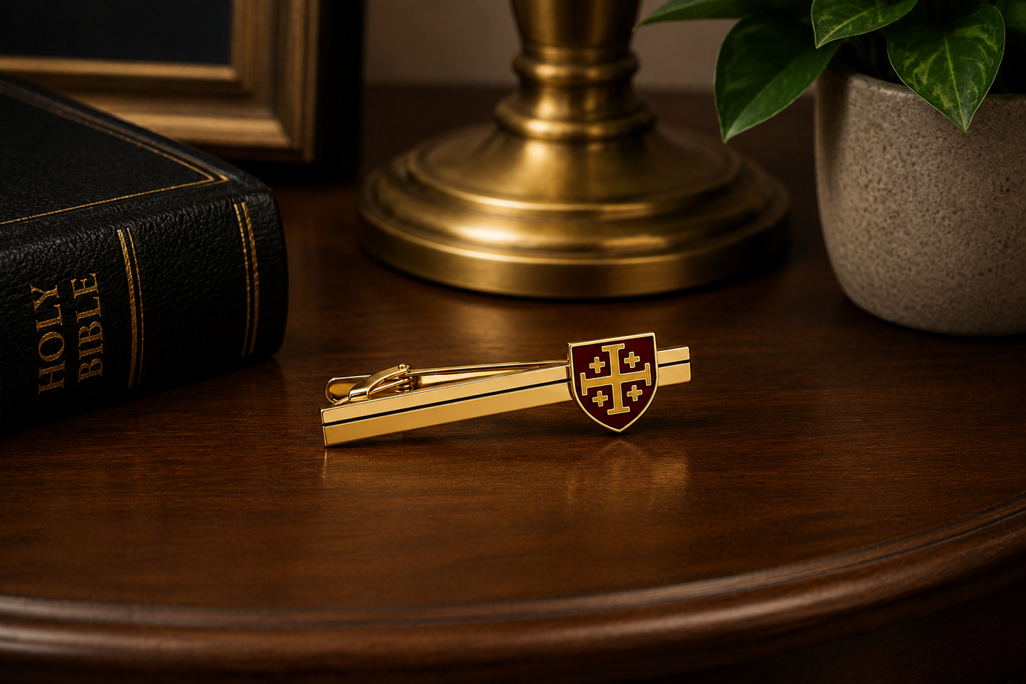 A gold tie clip featuring a small shield emblem with a red cross, displayed on a dark wooden table next to a Holy Bible, a brass candlestick base, and a potted plant.