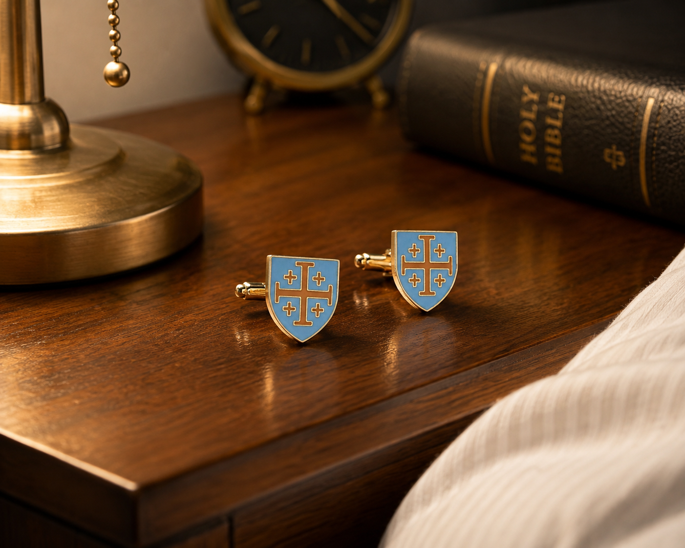 A pair of gold-tone cufflinks shaped like small blue shields with a gold cross, displayed on a dark wooden table next to a brass lamp base, a clock, a Holy Bible, and a white ribbed fabric.