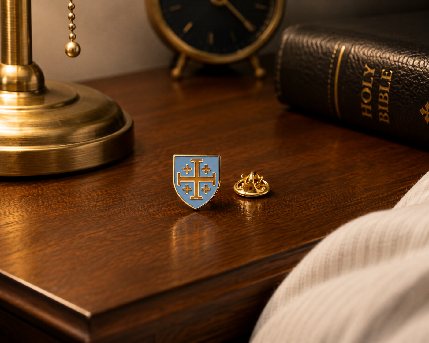 A small gold-toned lapel pin shaped like a shield with a blue background and gold cross detail, shown beside a matching gold pin on a wooden table. A brass lamp base, clock, Holy Bible, and draped fabric are visible in a styled indoor setting.