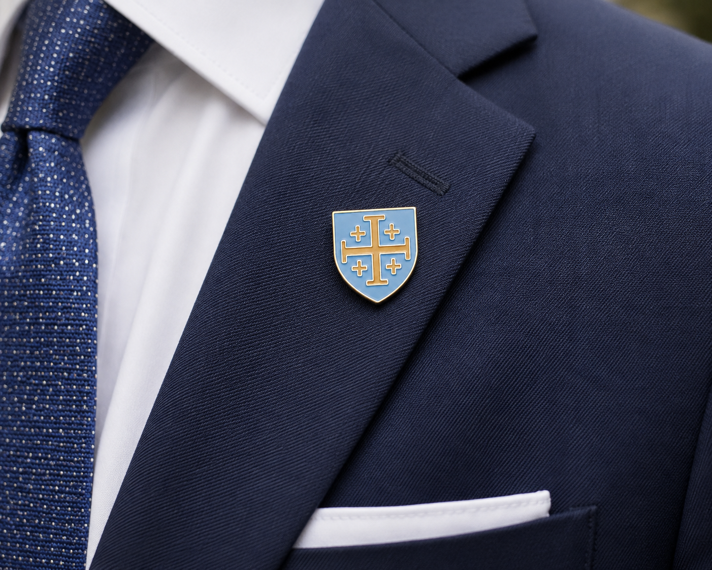 A close-up of a navy blue suit jacket with a white dress shirt, a blue patterned tie, and a white pocket square, featuring a small shield-shaped lapel pin with a gold cross and blue background on the chest.