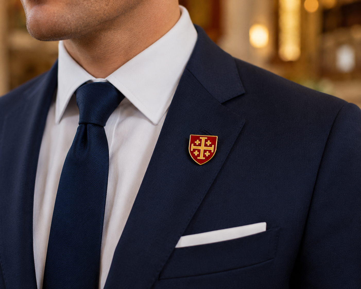 A close-up of a man wearing a navy blue suit jacket, white dress shirt, navy tie, and a small red and gold shield-shaped lapel pin on the chest.