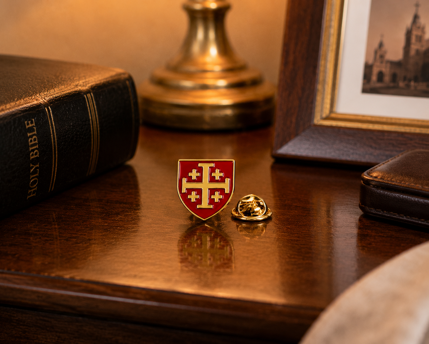 A small red and gold shield-shaped lapel pin with a cross motif sits on a polished wooden desk next to its gold-toned metal clasp. The scene includes a closed Holy Bible, a brass lamp base, and a framed picture in the background.