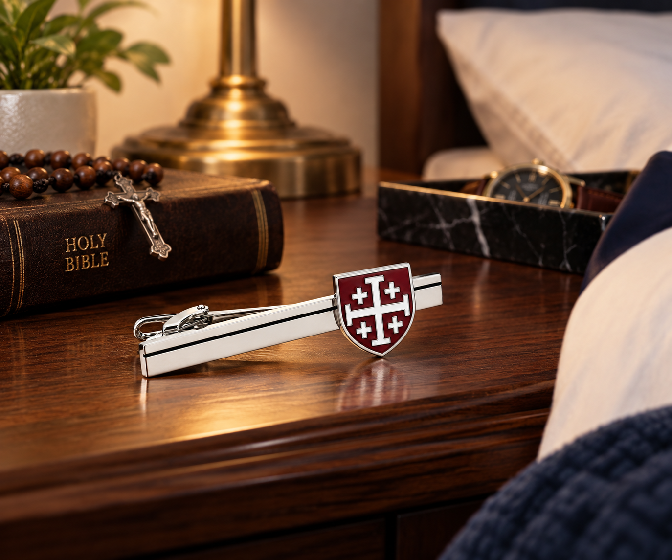A silver-toned tie bar with a shield-shaped red and white crest, displayed on a polished wooden bedside table next to a Holy Bible, a lamp, and a watch.