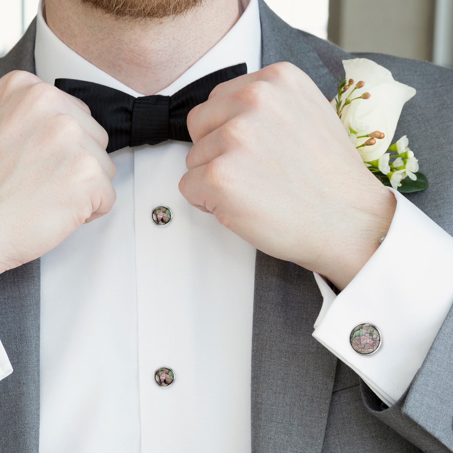 Man adjusting a black bow tie with a white shirt and gray suit jacket, wearing a white flower boutonniere.