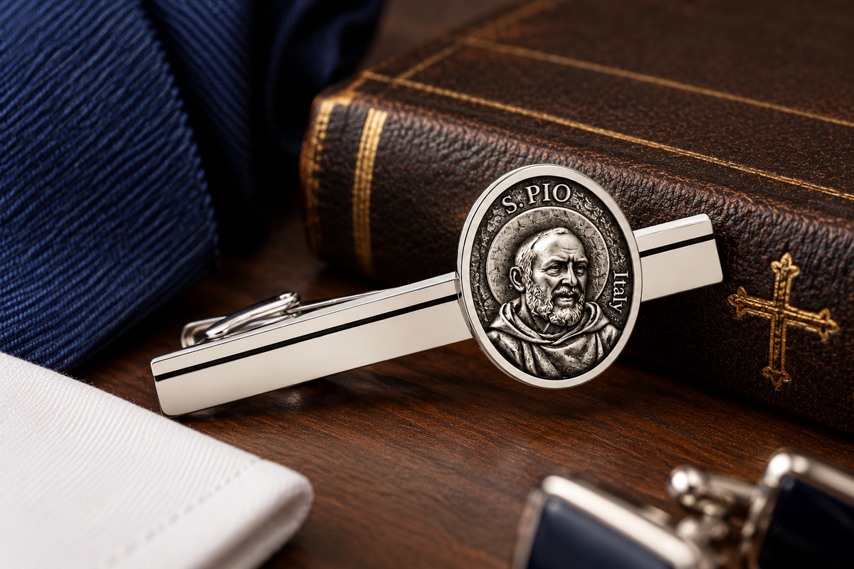 A silver-toned tie bar featuring an oval Saint Pio of Pietrelcina medal design, displayed on a dark wooden surface next to a navy blue tie, a brown leather-bound book, and a white pocket square.