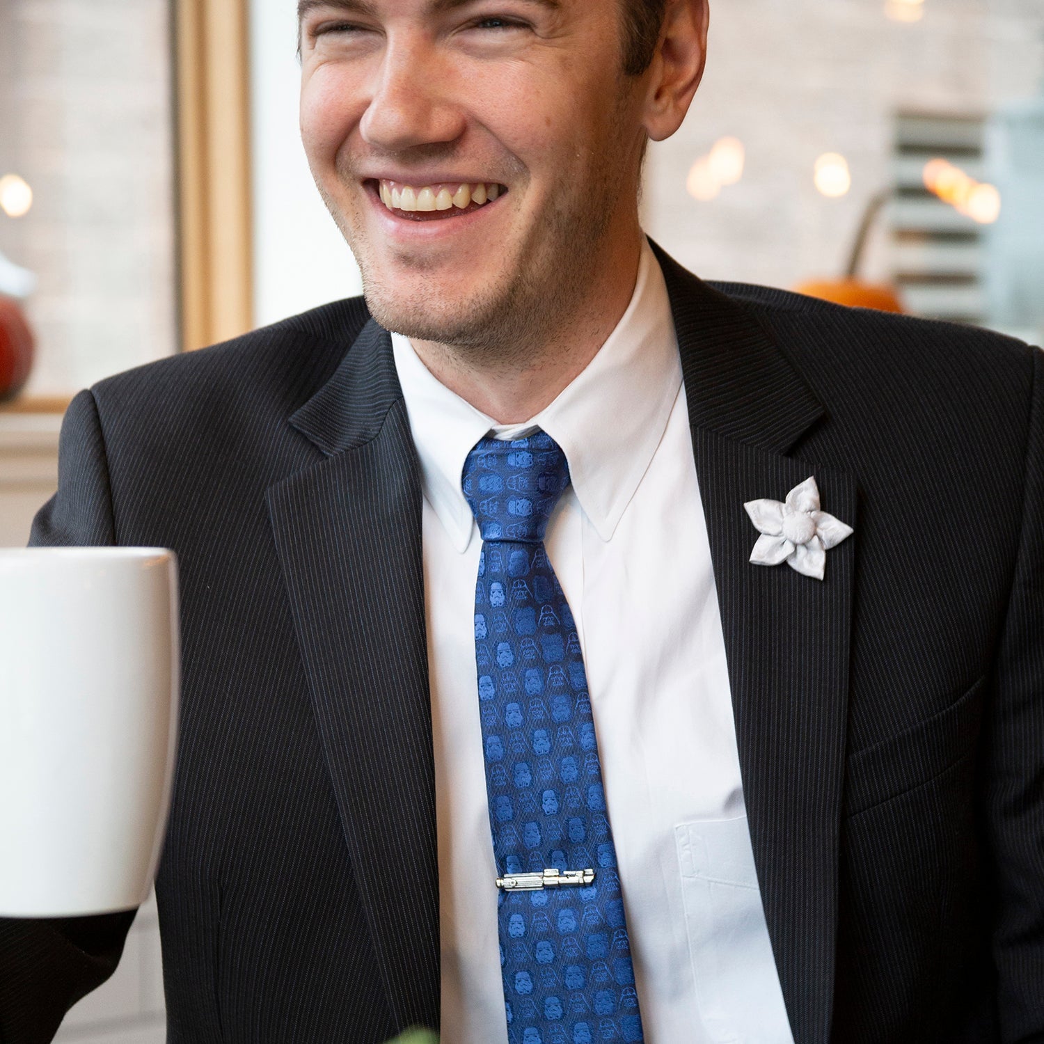Man in a suit with a blue tie and white flower pin, holding a white mug.