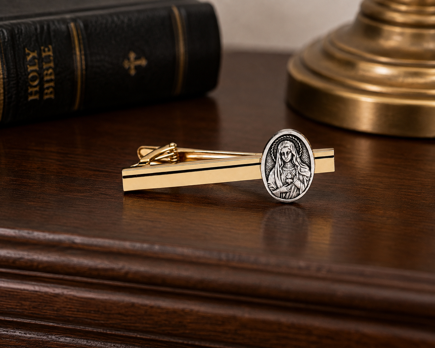 A gold-toned tie clip with a silver-toned oval medallion featuring a religious figure, displayed on a dark wooden surface with a Holy Bible and a brass lamp base in the background.