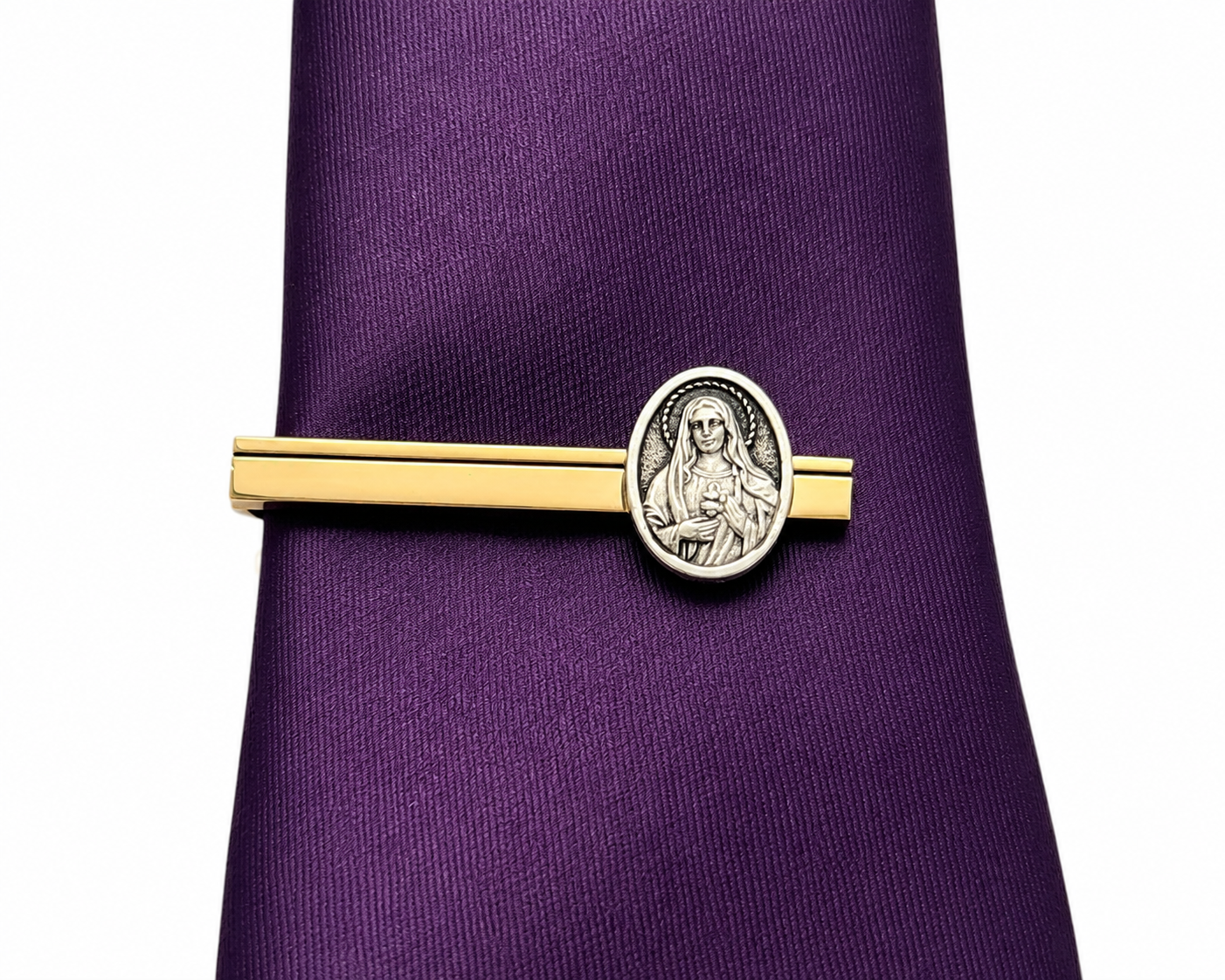 A gold and silver tie clip with an oval religious medallion, shown fastened to a purple silk necktie on a white background.
