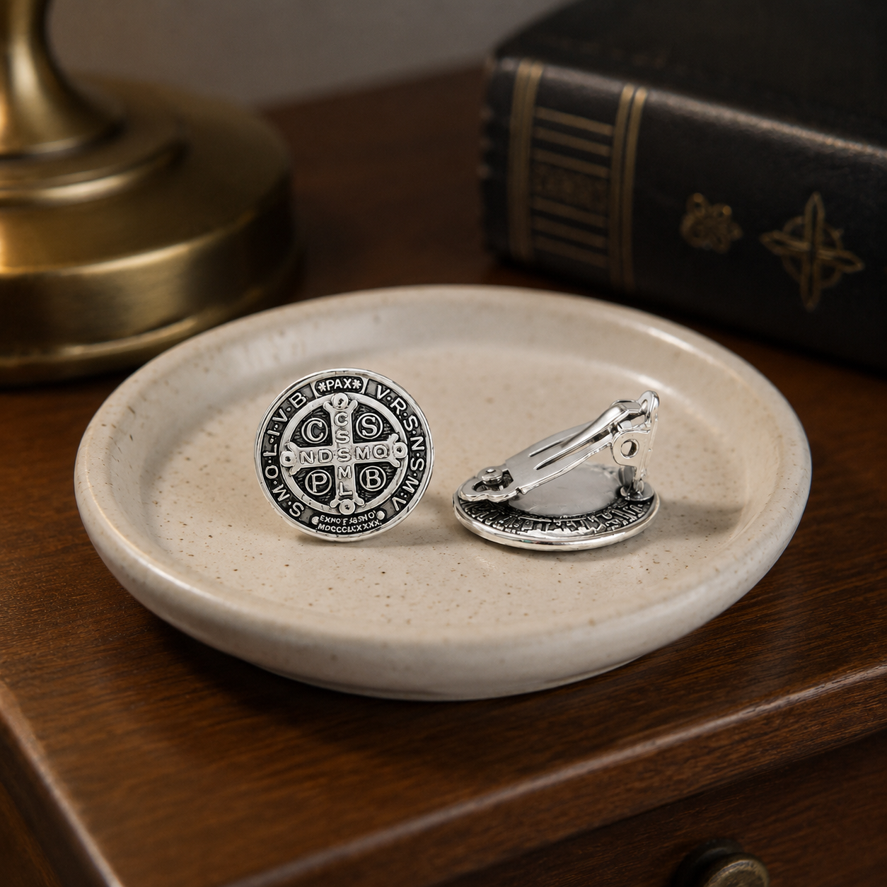 Silver Saint Benedict clip-on earrings resting in a ceramic jewelry dish on a wooden nightstand.