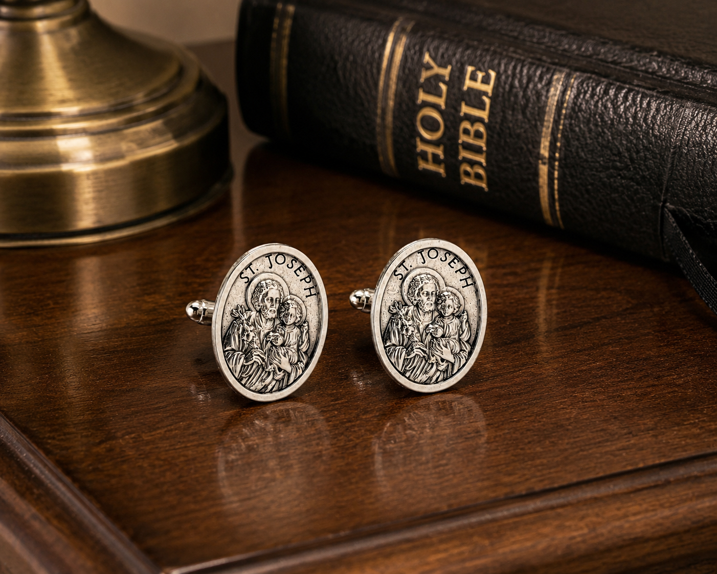 A pair of oval silver cufflinks featuring an engraved depiction of St. Joseph, displayed on a polished wooden table next to a Holy Bible and a brass lamp base.