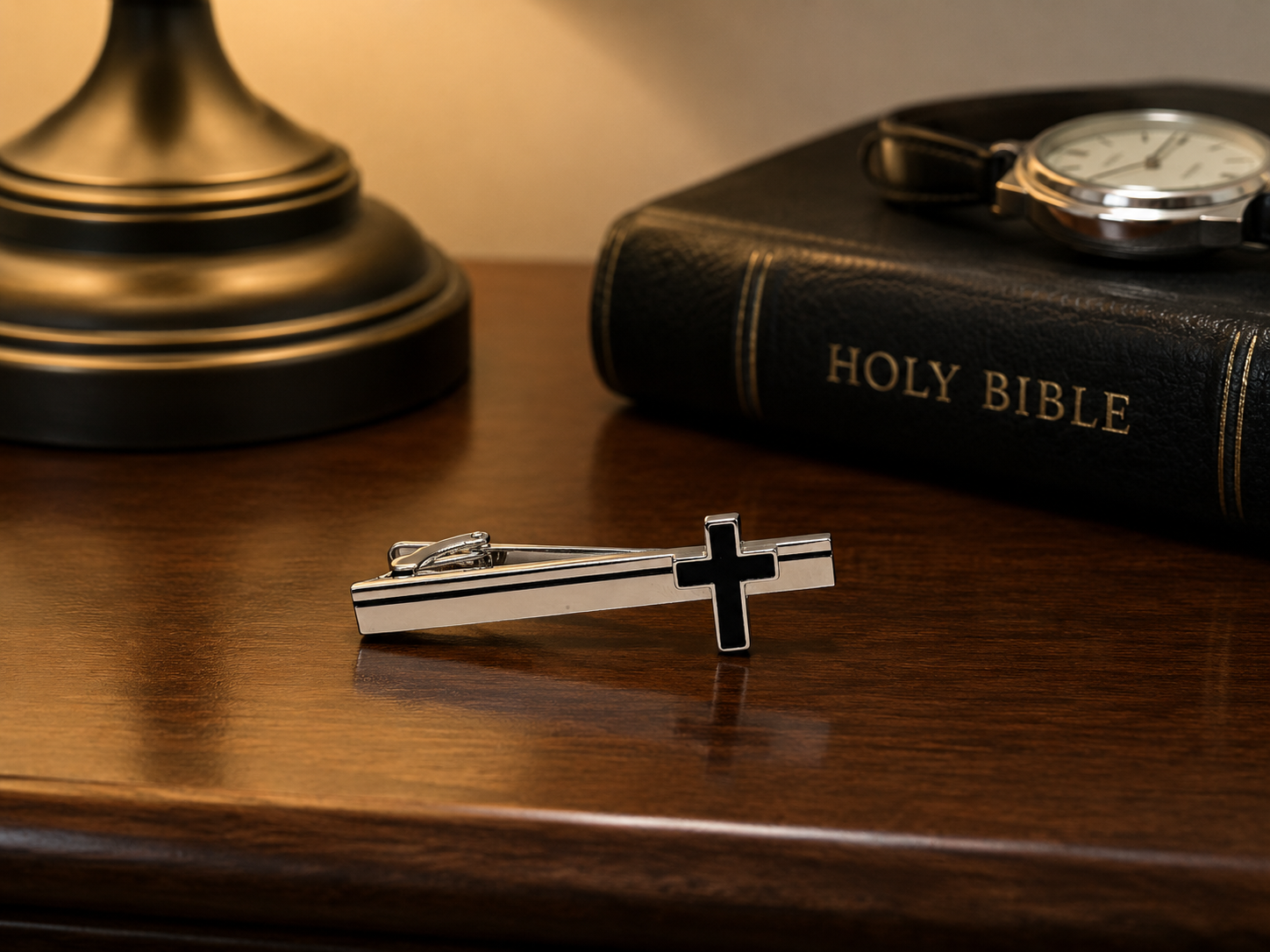 Silver tie clip with cross design on a wooden surface next to a Holy Bible and a lamp.