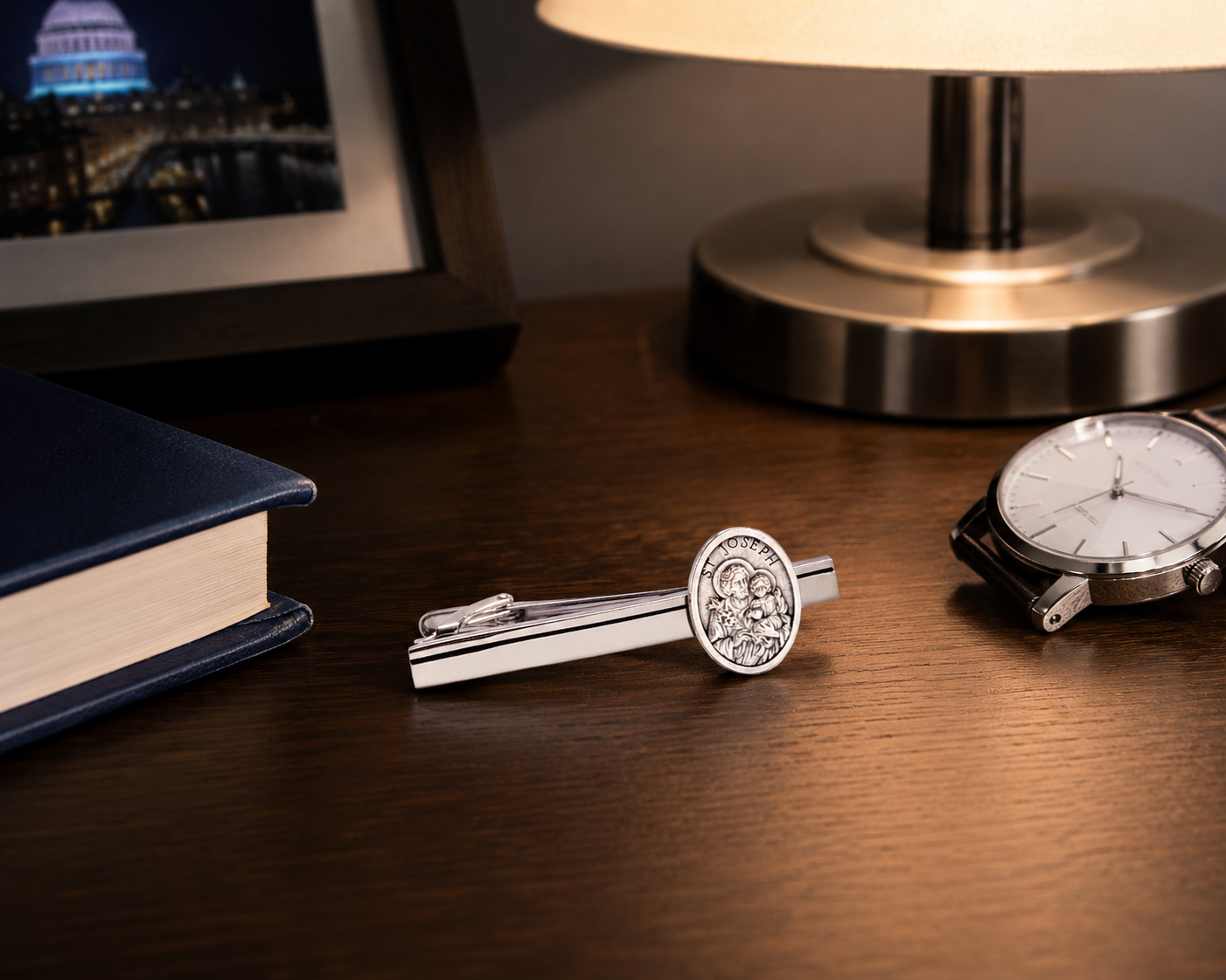 Silver tie clip with intricate design on a wooden surface with a book and lamp in the background.