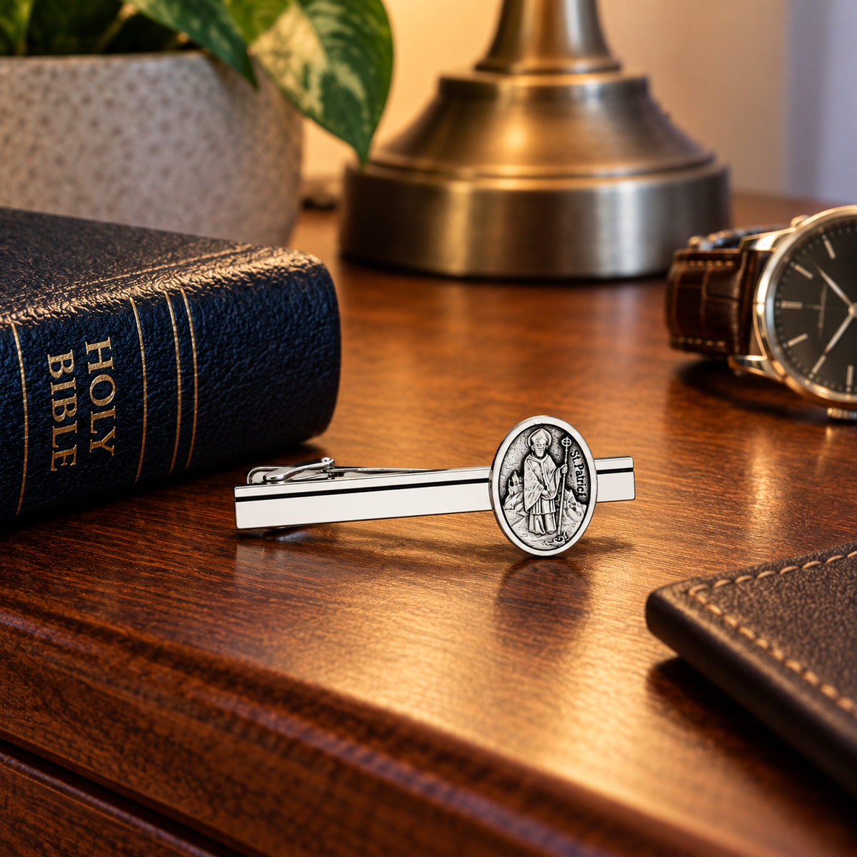 A silver-toned tie bar featuring an engraved circular medallion of Saint Patrick, displayed on a polished wooden desk alongside a Holy Bible, a watch, and a plant.