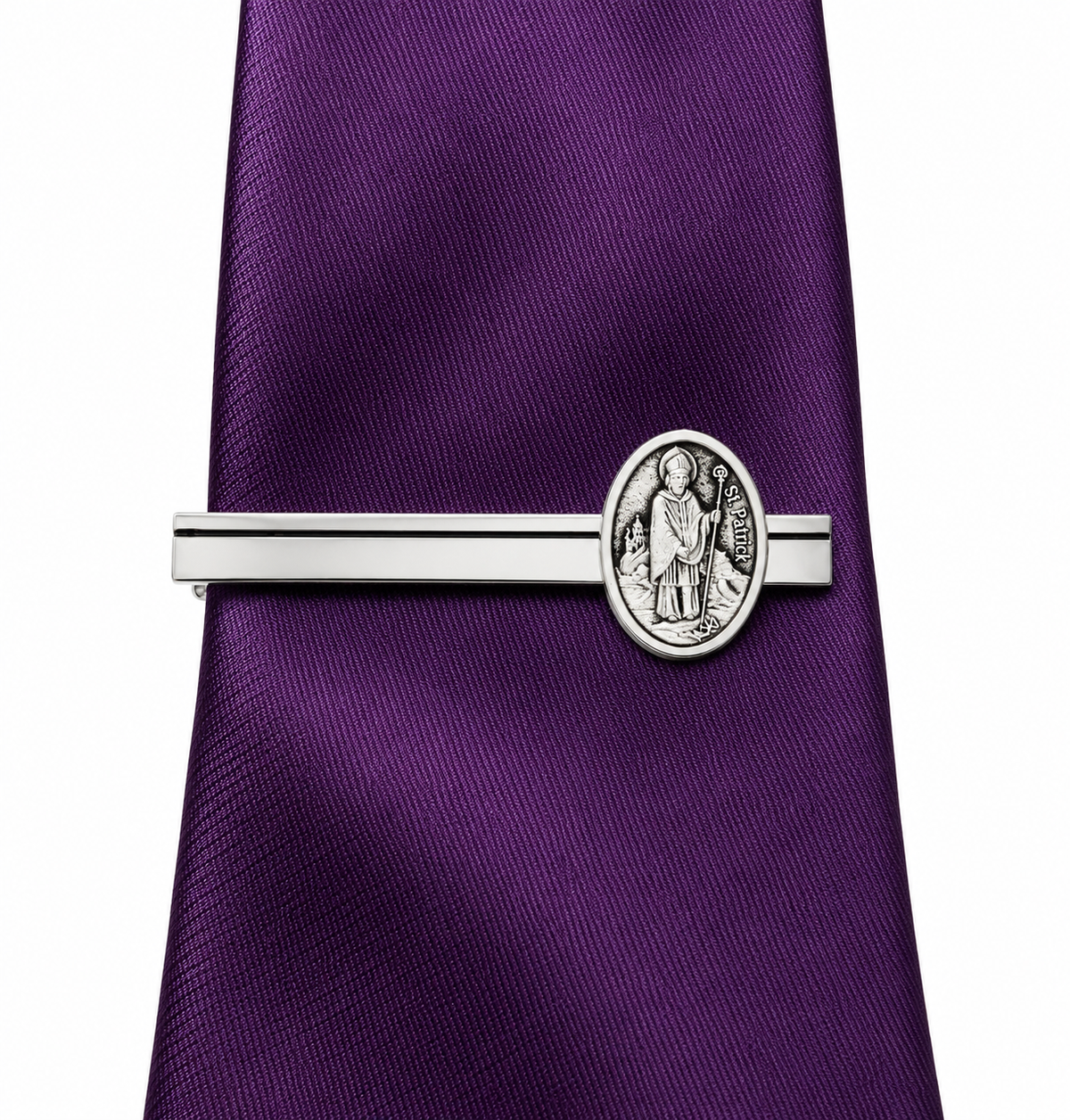 A silver-toned tie clip featuring an oval Saint Patrick medal, shown fastened to a purple textured silk tie on a white background.