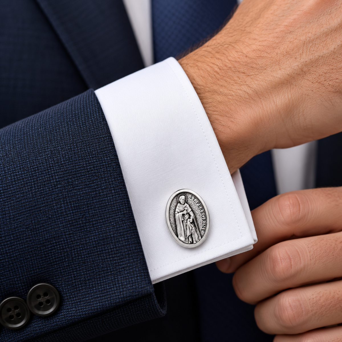 A silver oval cufflink featuring a religious saint design, shown fastened to a white dress shirt cuff worn under a navy blue suit jacket.