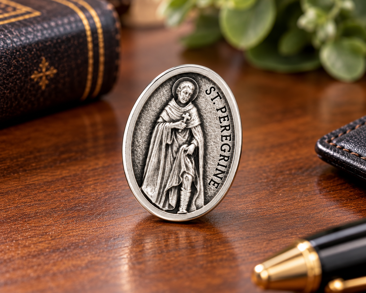 An oval silver-toned Saint Peregrine medal or ring clasp standing upright on a polished wooden desk, surrounded by a book, pen, and blurred greenery in the background.