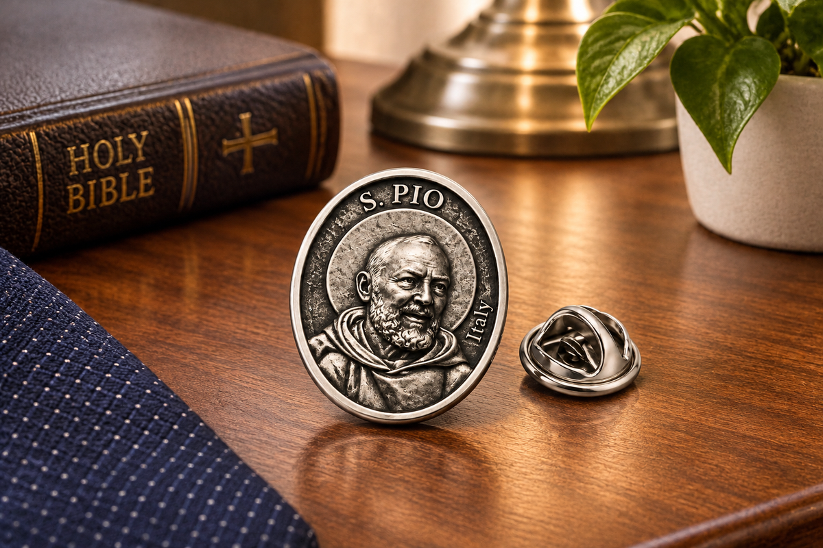 A silver-toned lapel pin shaped like a Saint Pio medallion, shown with its reverse side and pin clasp on a polished wooden tabletop. A dark leather Holy Bible, a folded navy blue textured fabric, and a potted green plant are styled in the background.