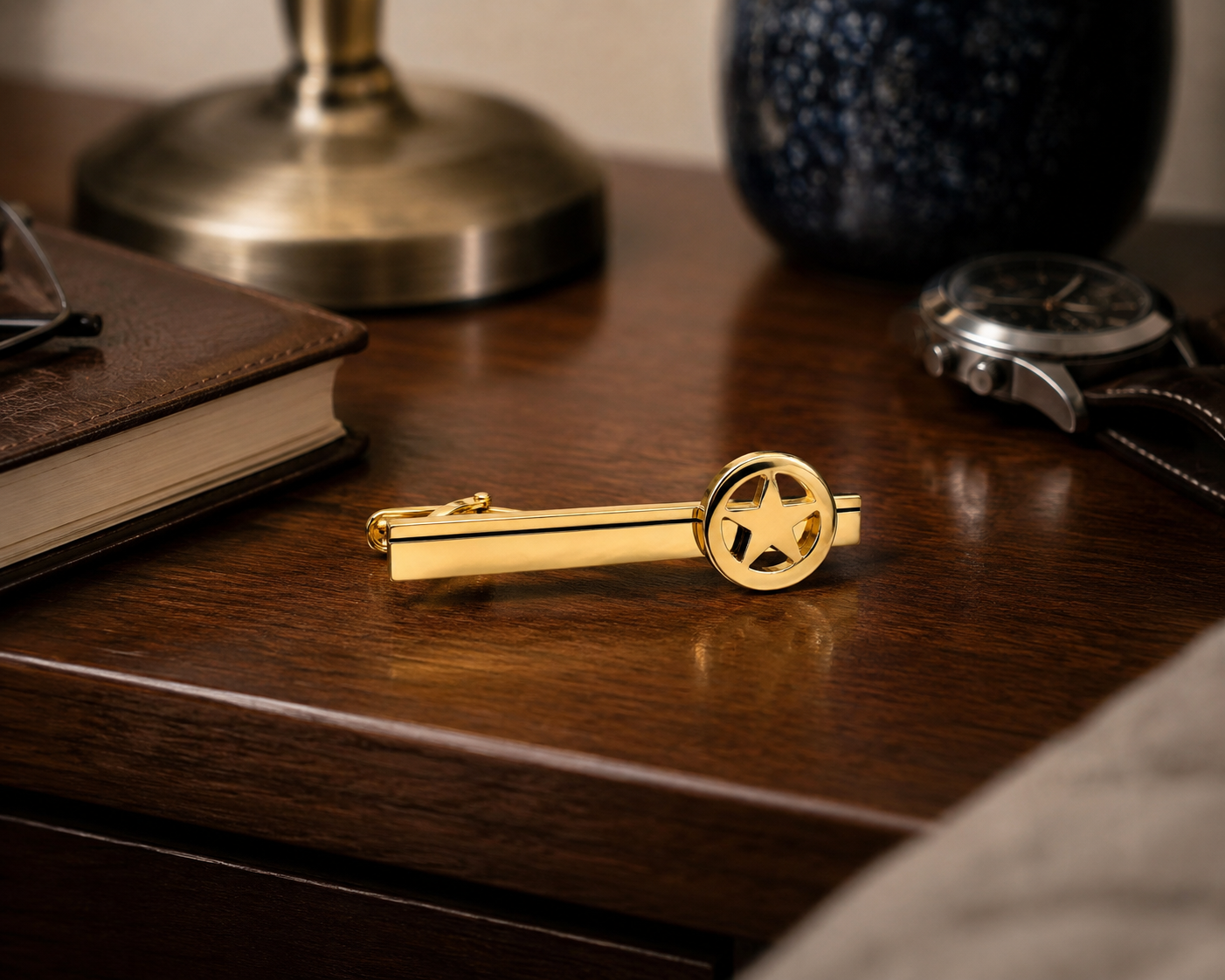 A gold tie bar with a star-shaped clip, resting on a dark wooden desk alongside a brown leather notebook, a watch, and a brass lamp base.