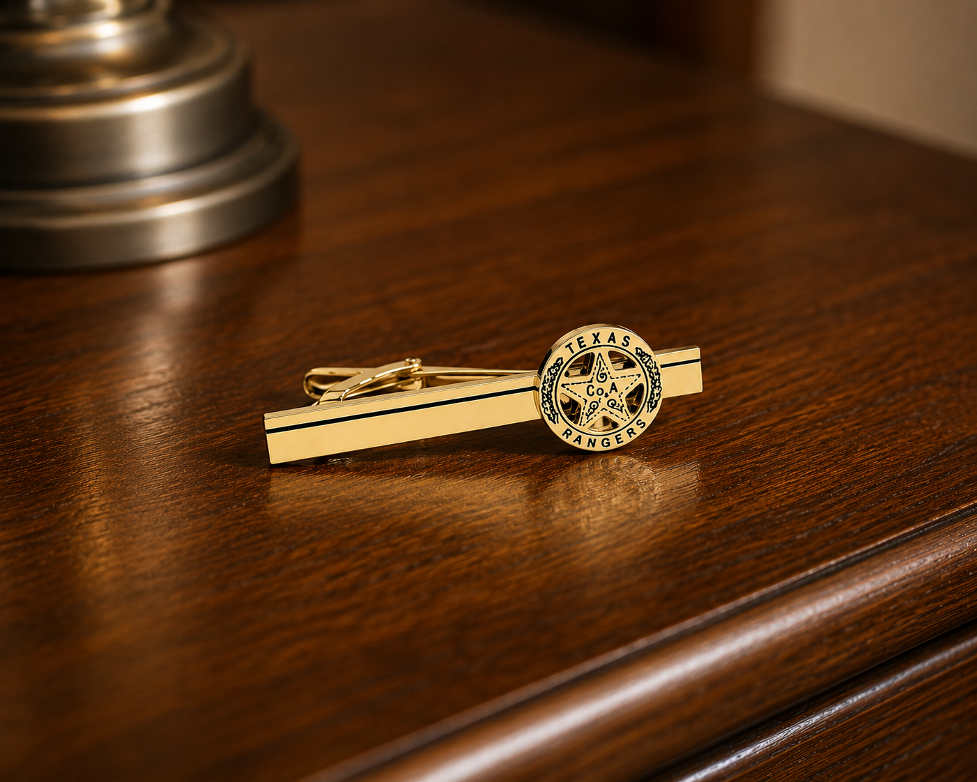 A gold tie bar featuring a circular Texas Rangers emblem, resting on a polished dark wood surface.