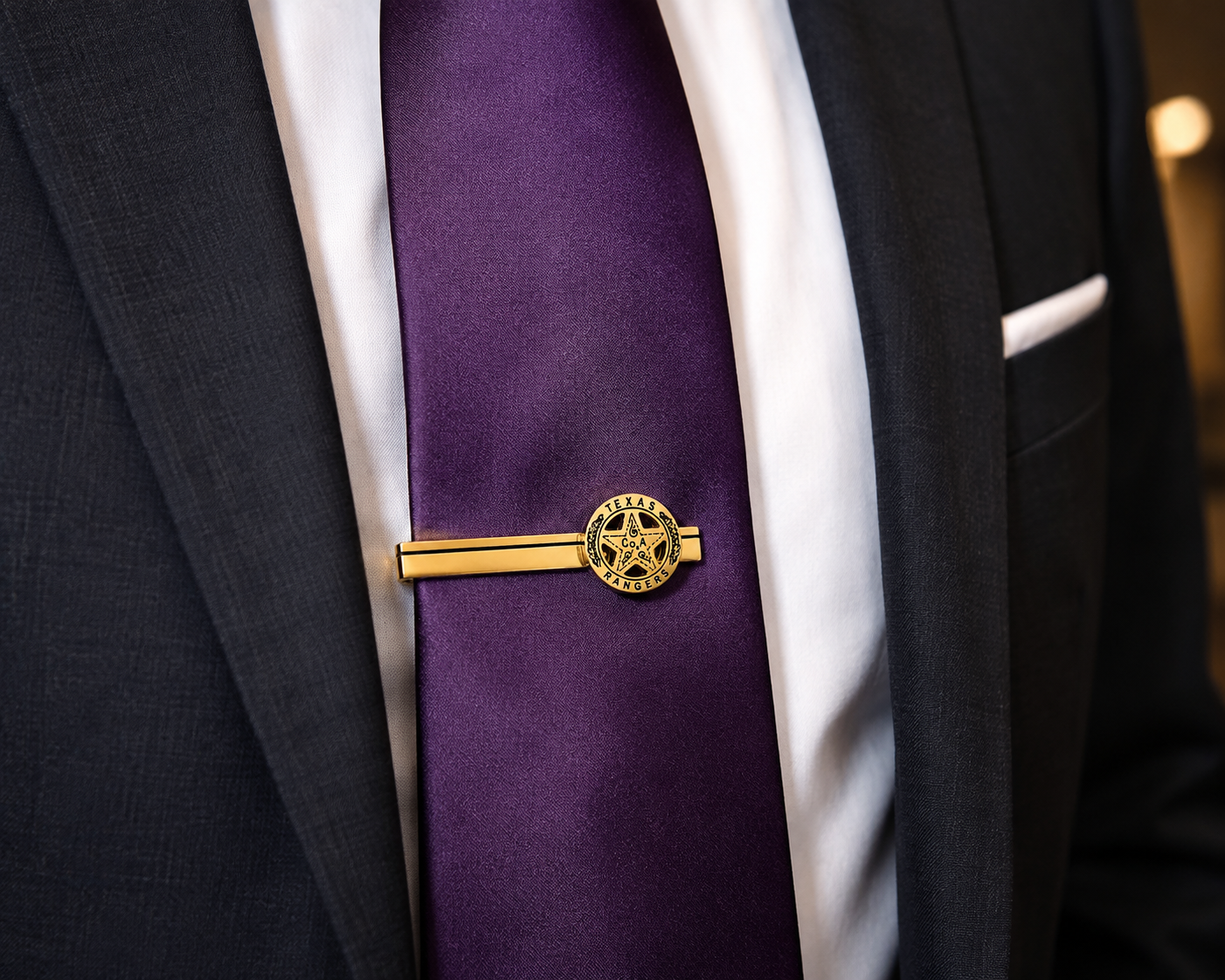 A gold metal tie bar with an engraved star emblem, holding a purple necktie on a white dress shirt and dark suit jacket.