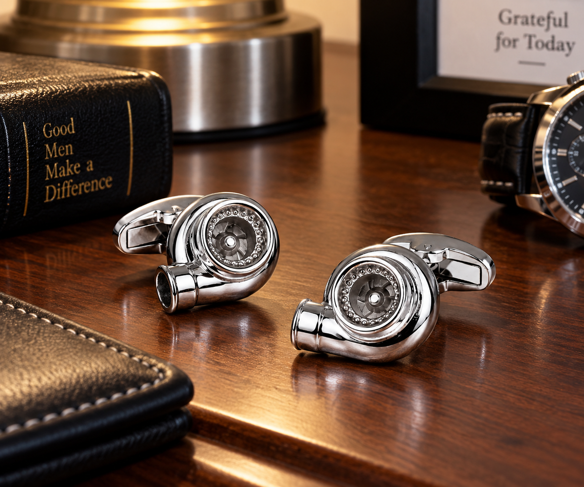 Two silver metallic cufflinks shaped like turbochargets, displayed on a polished wooden desk alongside other lifestyle props including a leather wallet, a watch, and a Bible.