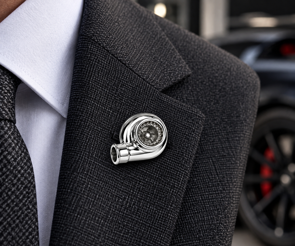 A silver metallic lapel pin shaped like a stylized swirl or spiral, worn on the lapel of a dark gray textured suit jacket over a white collared shirt.