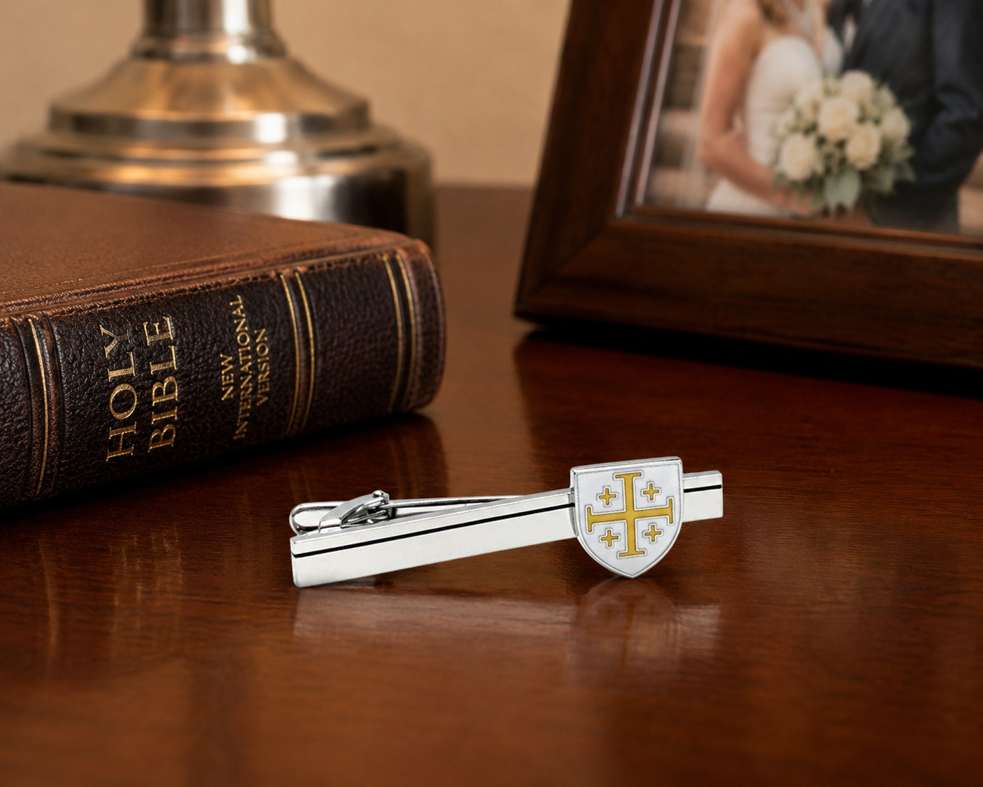 A silver-toned tie clip with a shield emblem featuring a gold cross and four gold stars, resting on a polished wooden table next to a brown leather Holy Bible and a framed photo.